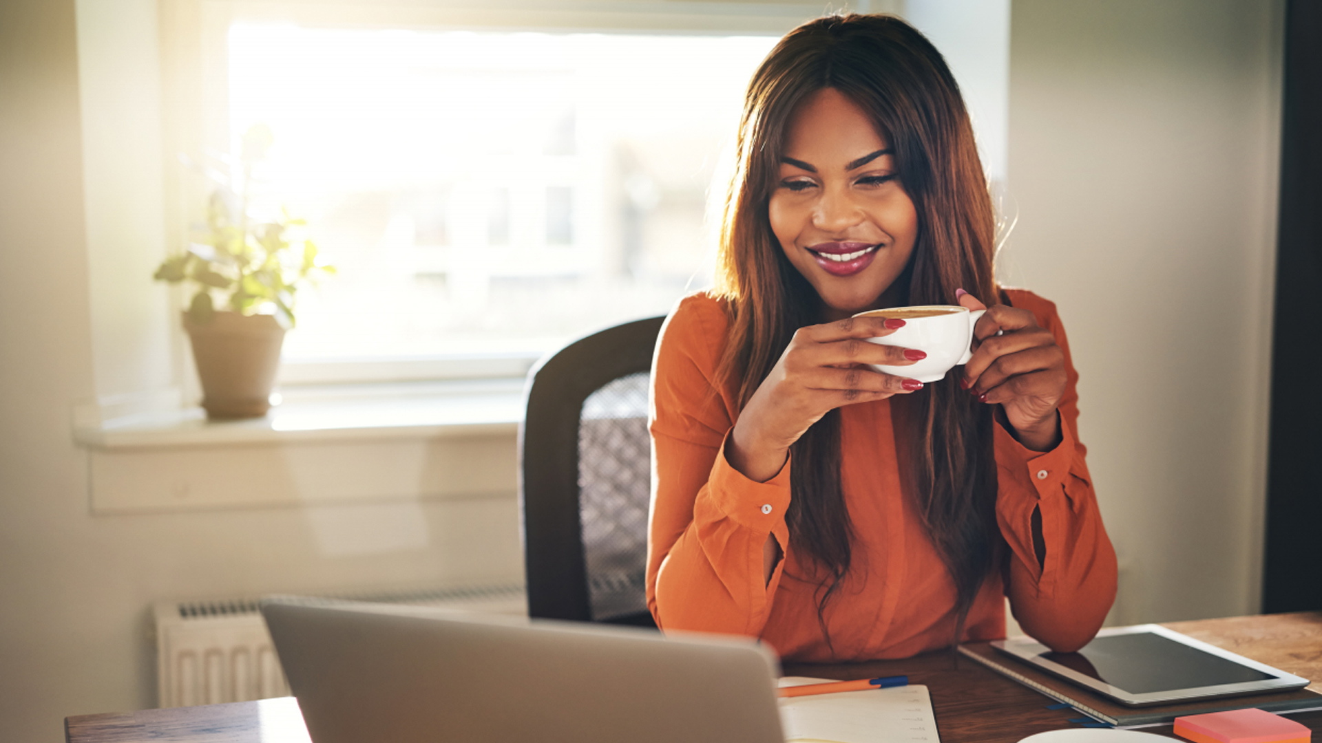 home office ideas woman in orange blouse sat at desk looking at laptop, holding a mug